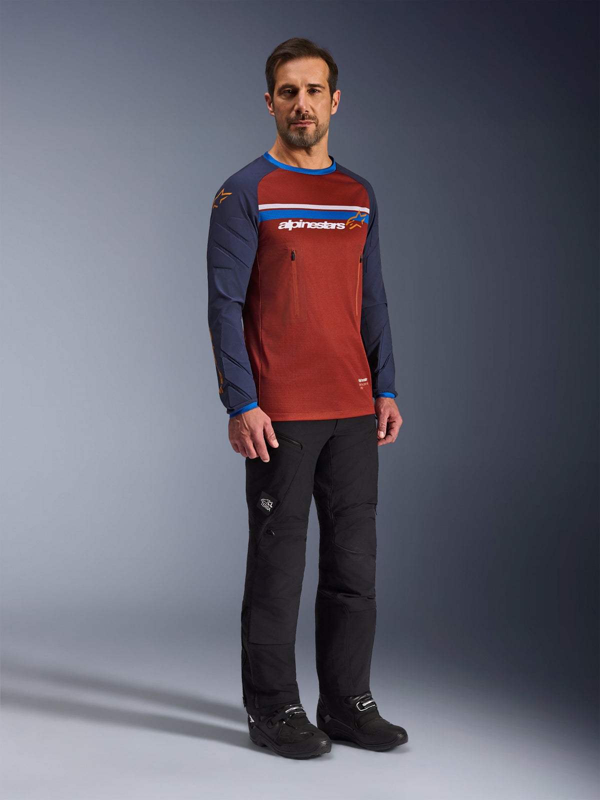 Mężczyzna w koszulce Alpinestars Maxdura Dual Jersey w kolorach Rust Brown i Mood Indigo, siatkowy tułów i wyściełane rękawy, czarne spodnie i buty motocyklowe, odzież motocyklowa adventure