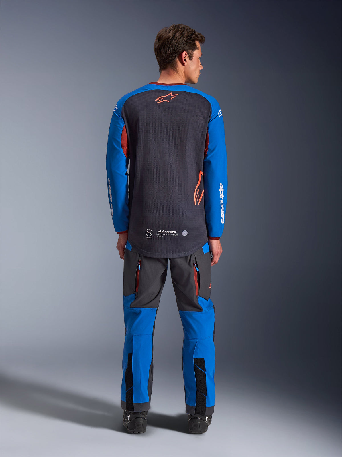Koszulka Alpinestars Maxdura Dual w kolorze ciemnoszarym i niebieskim, widok z tyłu pokazujący siatkowy tors i pomarańczowe logo z gwiazdą, niebieskie rękawy z białym brandingiem, zestawiona z dopasowanymi szaro-niebieskimi spodniami adventure, wysokiej klasy odzież do jazdy typu dual-sport