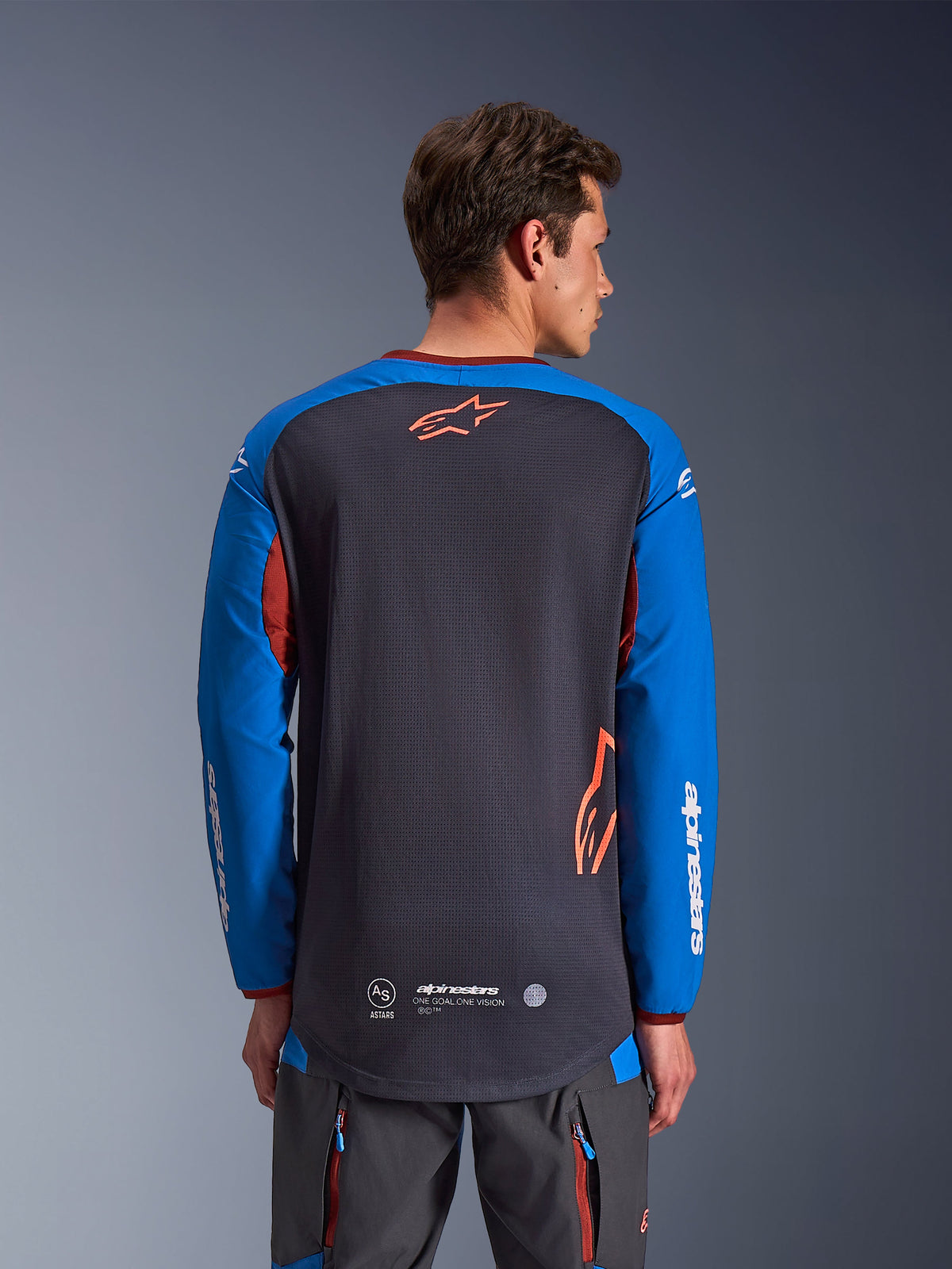 Alpinestars Maxdura Dual Jersey w kolorze ciemnoszarym i królewskim niebieskim, widok od tyłu na modelu, siateczkowa konstrukcja tułowia z pomarańczowym logo Astars, długie rękawy z białym brandingiem, bordowy kołnierzyk i mankiety, zestaw z szarymi spodniami technicznymi, odzież motocyklowa dual-sport i adventure