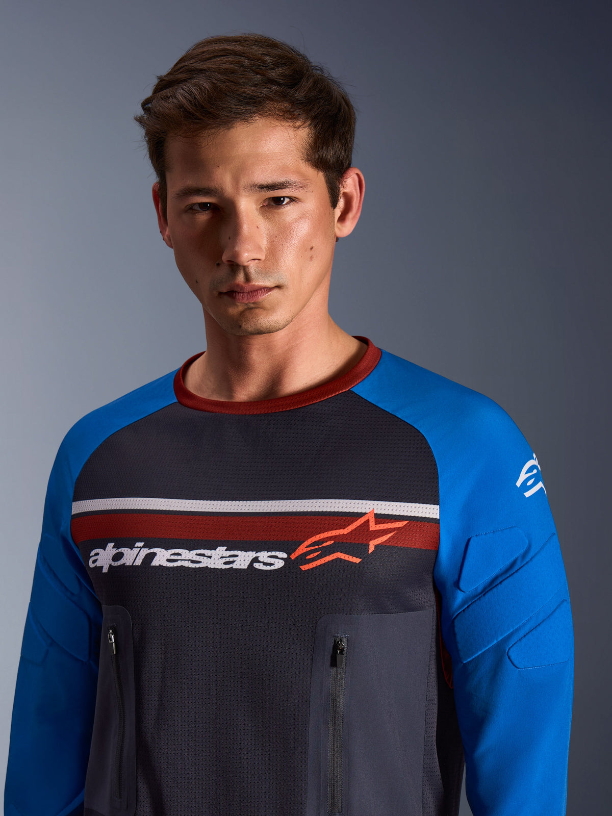 Alpinestars Maxdura Dual Jersey, ciemnoszara i niebieska (Royal Blue), prezentowana przez modela, z siateczkowym tułowiem, niebieskimi rękawami ze zintegrowaną wyściółką oraz pionowymi kieszeniami na klatce piersiowej zapinanymi na zamek i logo Alpinestars.