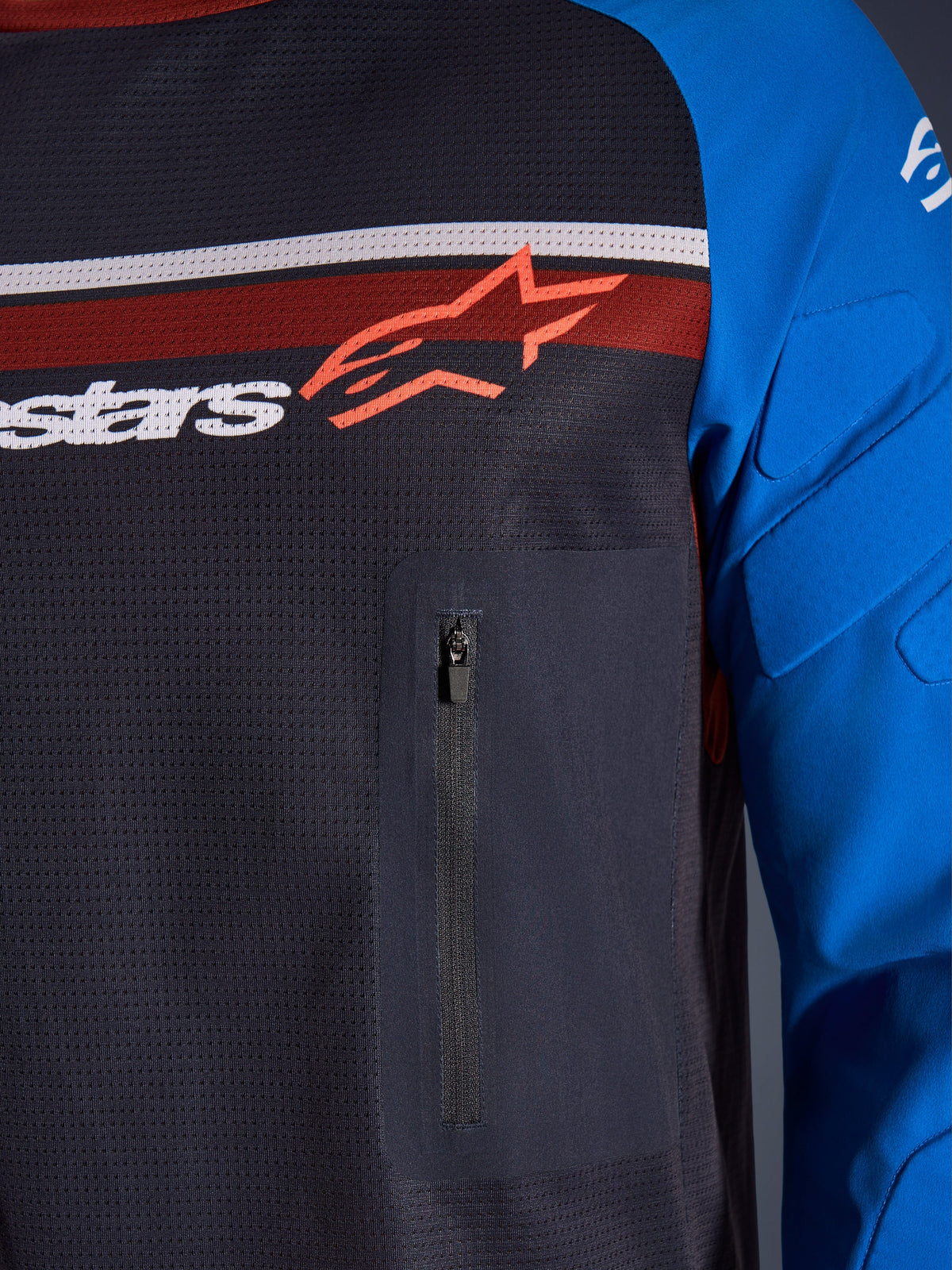 Alpinestars Maxdura Dual Jersey, zbliżenie na klatkę piersiową i rękaw, ciemnoszary i niebieski, zapinana na zamek kieszeń na piersi, oddychający siatkowy tułów, zintegrowane ochraniacze na rękawach, logo Alpinestars na tle czerwonych i białych pasów