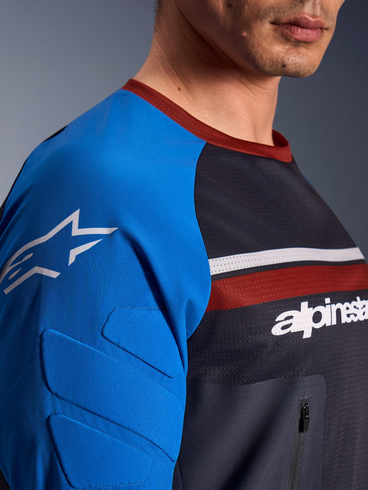 Alpinestars Maxdura Dual Jersey, koszulka motocyklowa, ciemnoszara i niebieska, widok z bliska na kieszeń na klatce piersiowej zapinaną na zamek, siatkowy materiał i zintegrowane ochraniacze na rękawach z logo gwiazdy