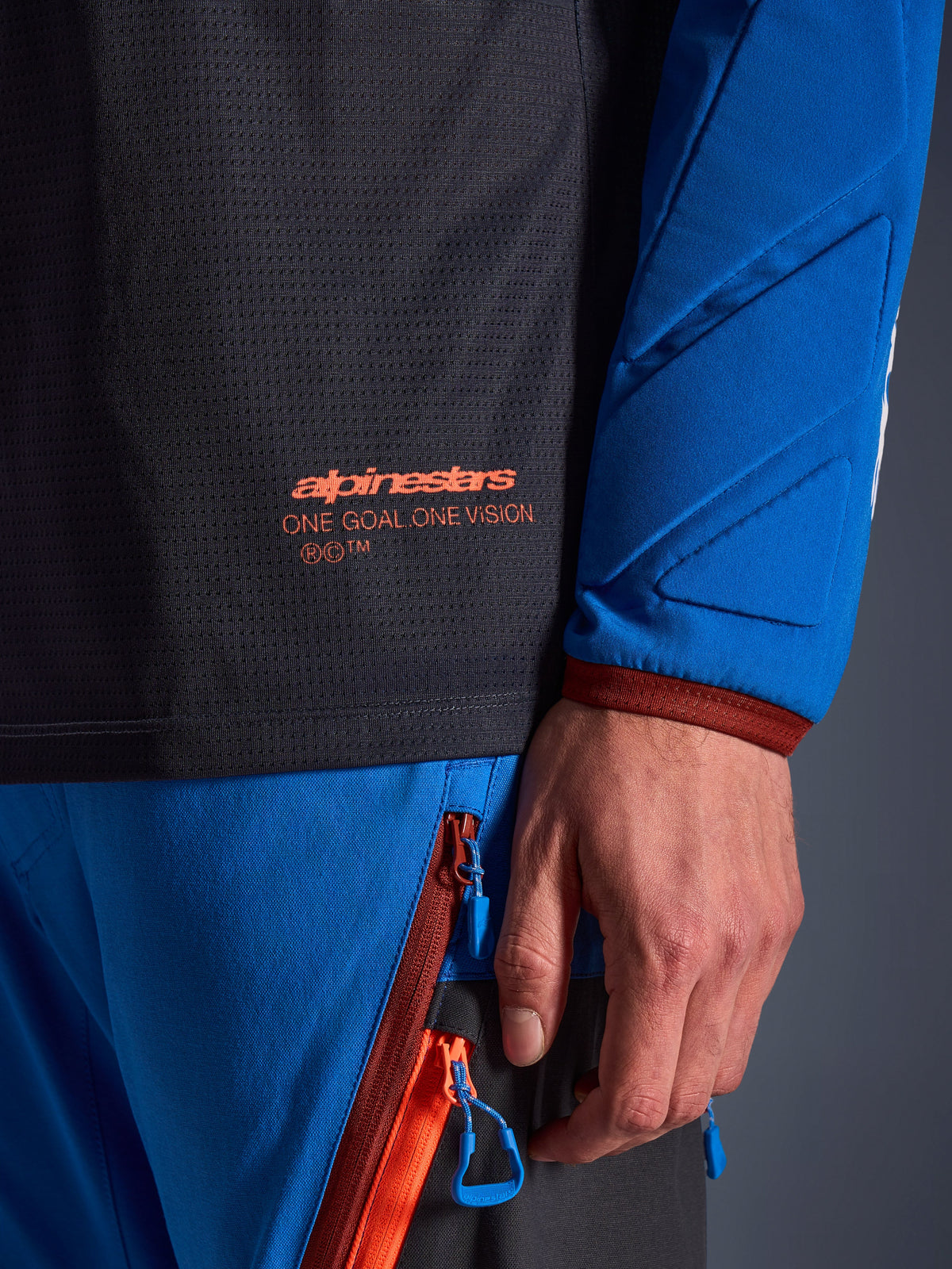 Zbliżenie na koszulkę Alpinestars Maxdura Dual w kolorze ciemnoszarym i niebieskim, pomarańczowe logo i napis „One Goal One Vision” na siatkowym tułowiu, wyściełany rękaw ze wzmocnionymi panelami geometrycznymi, detal technicznej odzieży adventure