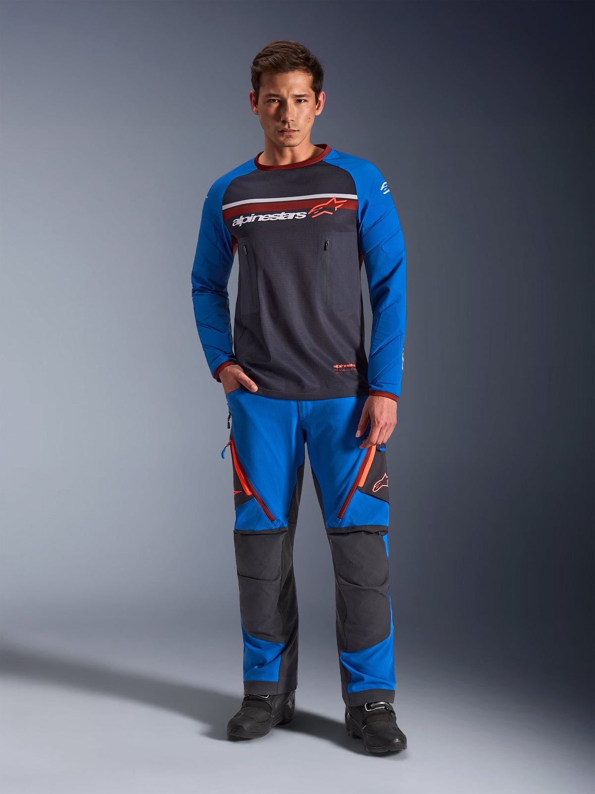 Alpinestars Maxdura Dual Jersey, koszulka motocyklowa, ciemnoszara i królewski błękit, tułów z pełnej siatki, wyściełane rękawy i kieszenie na piersi na zamek, białe i pomarańczowe logo Alpinestars, noszona z dopasowanymi spodniami adventure i czarnymi butami
