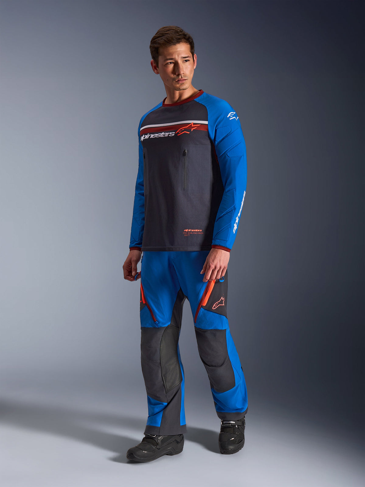 Alpinestars Maxdura Dual Jersey, ciemnoszara i niebieska, koszulka motocyklowa adventure z siatkowym tułowiem i niebieskimi rękawami, noszona przez modela wraz z pasującymi spodniami terenowymi, zapinane na zamek kieszenie na piersi, logo Alpinestars