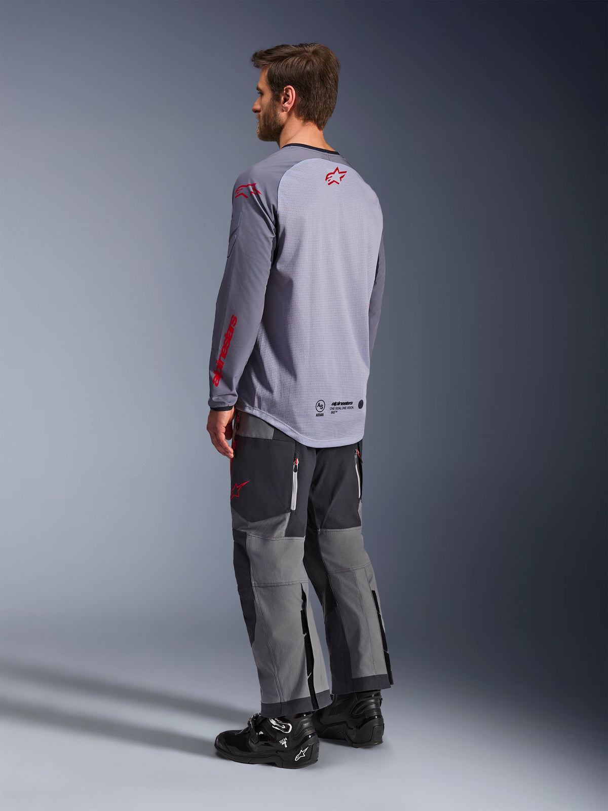 Man wearing Alpinestars Maxdura Dual Jersey, light gray with red Alpinestars logos, back view showing mesh torso and long sleeves, paired with charcoal technical pants and black motorcycle boots