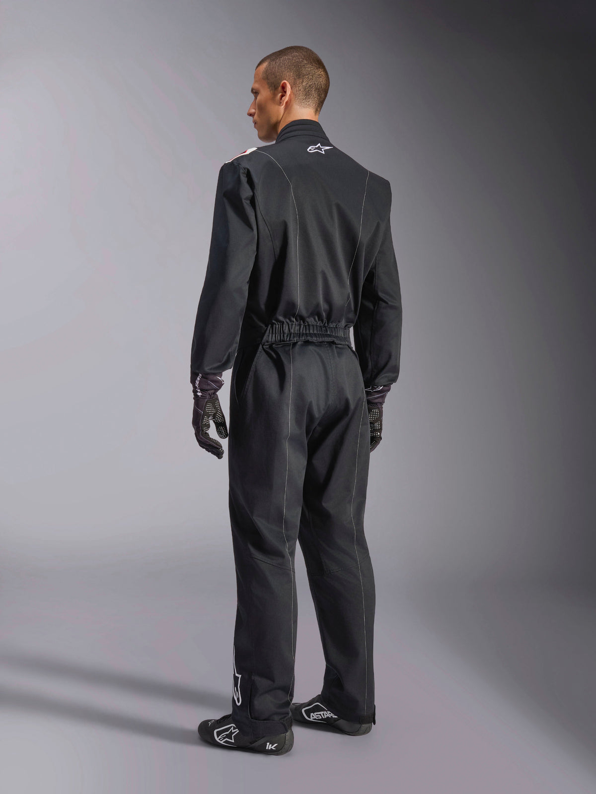 Alpinestars Kart Indoor V2 Suit, Black one-piece karting suit shown from a back three-quarter angle, featuring an elastic waist and a white logo on the neck, worn with black racing gloves and shoes, durable design for recreational karting
