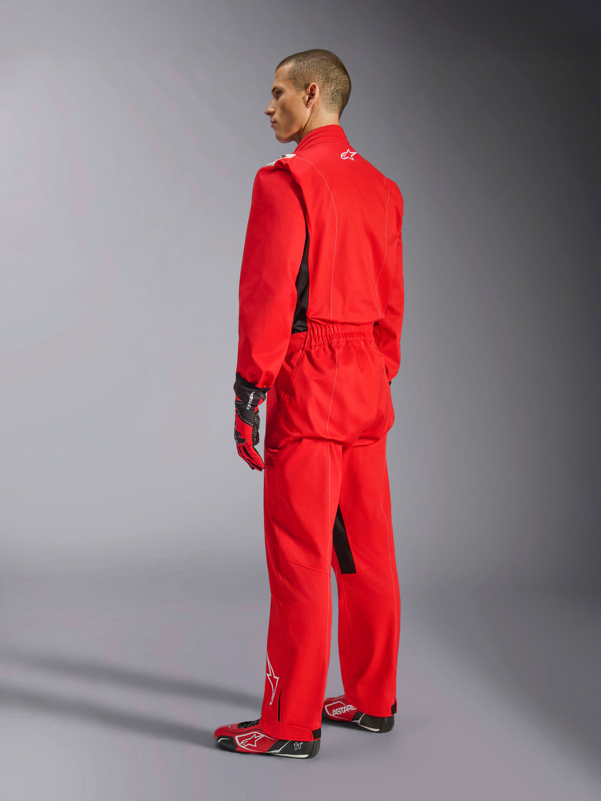 Alpinestars Kart Indoor V2 Suit, czerwony kombinezon kartingowy, widok z tyłu pod kątem trzech czwartych na modelu, zestawiony z pasującymi czerwonymi rękawicami i butami wyścigowymi, jednowarstwowa konstrukcja z kepru polibawełnianego, profesjonalny sprzęt motorsportowy do kartingu halowego.