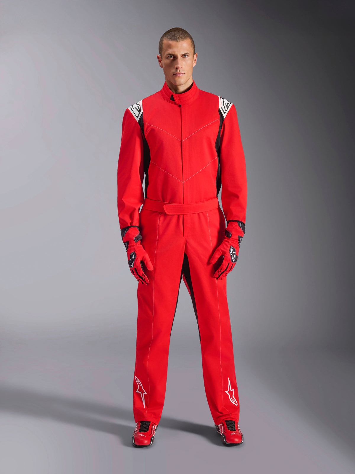 Alpinestars Kart Indoor V2 Suit, czerwony, jednoczęściowy kombinezon kartingowy noszony z pasującymi czerwonymi rękawicami i butami wyścigowymi, białe logo na ramionach i nogawkach, czarne akcenty, nowoczesny krój do wyścigów rekreacyjnych