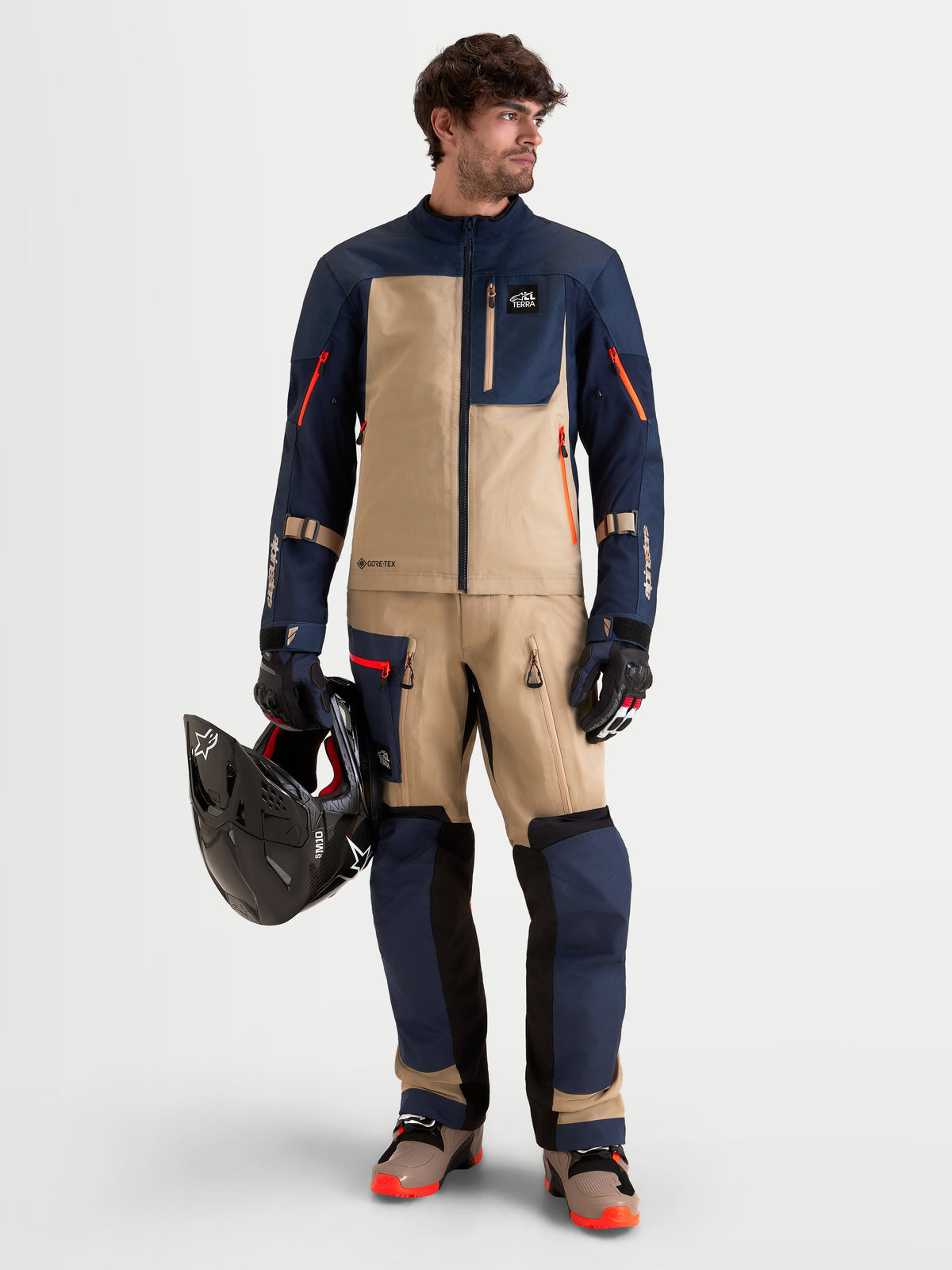 Alpinestars Arenis Gore-Tex Gear Set, męski zestaw motocyklowy adventure, granatowy, beżowy i czarny, pełny widok z przodu na modelu w pasującej odzieży trzymającym kask, wodoodporna odzież motoryzacyjna All-Terrain z certyfikatem AA