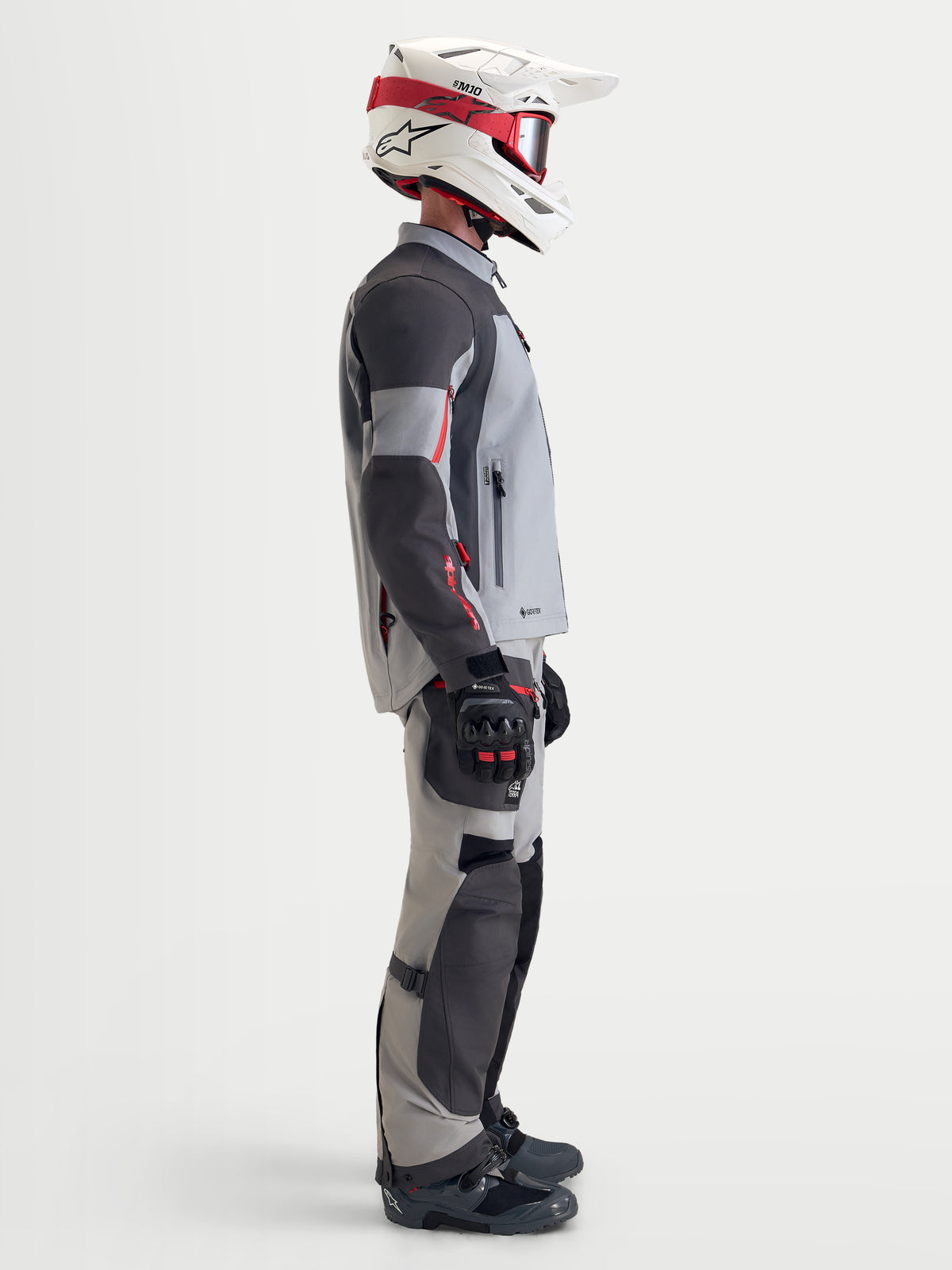 Alpinestars Arenis Gore-Tex kurtka motocyklowa męska turystyczna w kolorze jasnoszarym, ciemnoszarym i czarnym, profilowy widok motocyklisty w wodoodpornej kurtce tekstylnej z pasującym wyposażeniem i biało-czerwonym kaskem