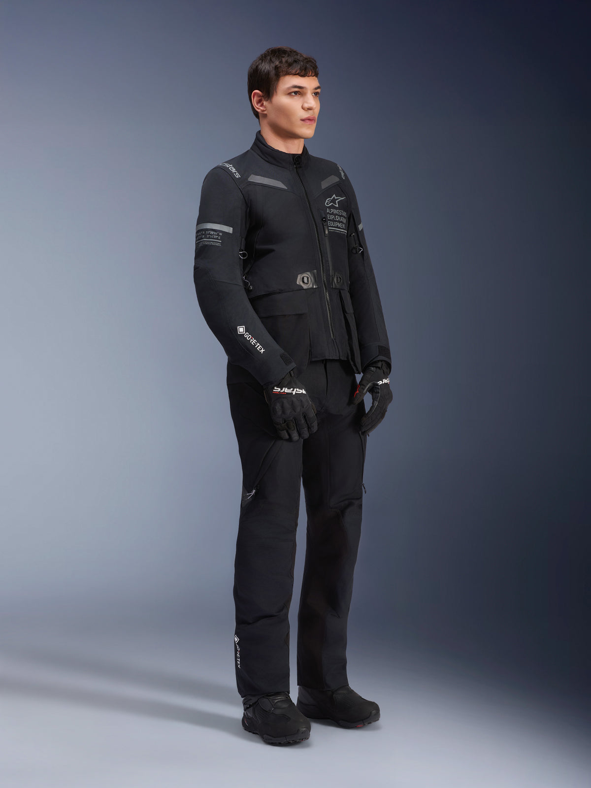 Spodnie Alpinestars ST-7 2L GORE-TEX, noszone przez modela z dopasowaną kurtką ST-7, czarny motocyklowy strój typu adventure touring, widoczne logo GORE-TEX, kieszenie cargo, czarne rękawice i buty, zdjęcie studyjne na szarym tle