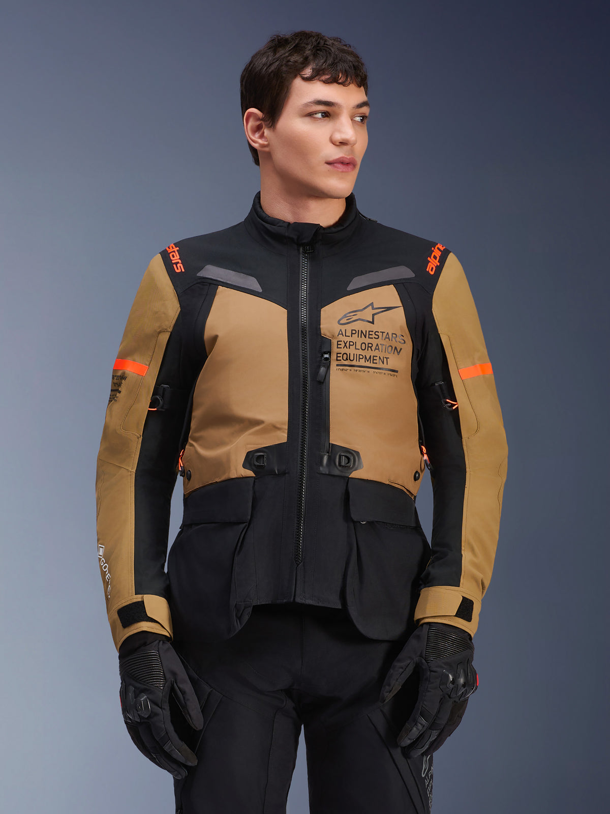 Kurtka Alpinestars ST-7 2L GORE-TEX, motocyklowa kurtka turystyczna adventure, kolor Gobi Brown i czarny z pomarańczowymi akcentami, prezentowana przez modela, z brandingiem Exploration Equipment i technologią GORE-TEX, wytrzymała wodoodporna odzież motocyklowa.