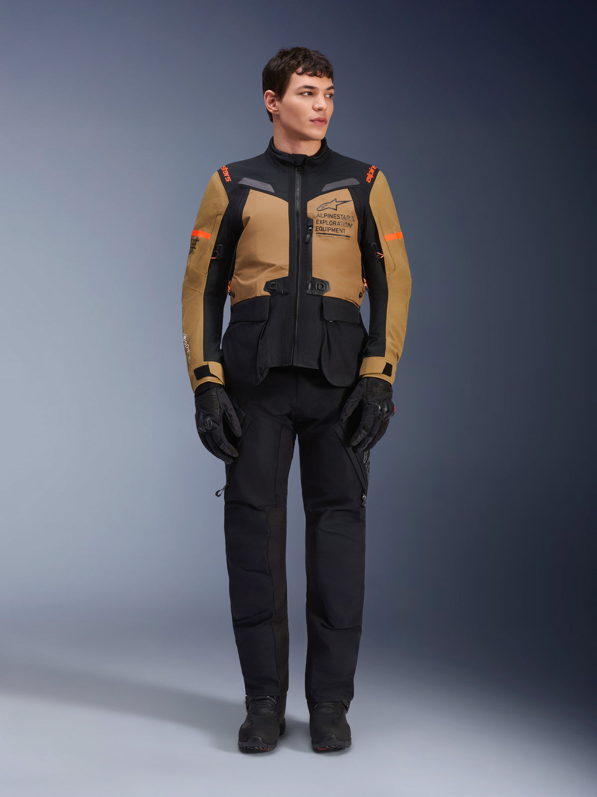 Męski model w kurtce Alpinestars ST-7 2L GORE-TEX, kolor Gobi Brown i czarny, odzież motocyklowa adventure z dopasowanymi czarnymi spodniami i rękawicami, logo Alpinestars Exploration Equipment na klatce piersiowej, funkcjonalna wodoodporna konstrukcja do turystyki szosowej i terenowej