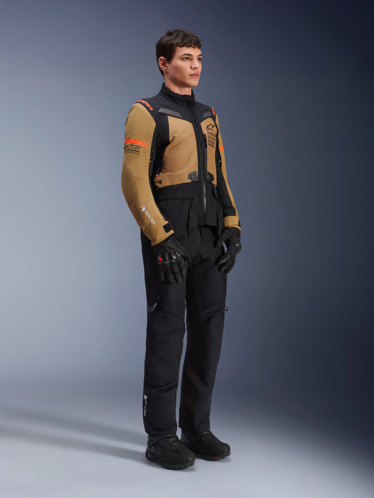 Kurtka Alpinestars ST-7 2L GORE-TEX, kurtka motocyklowa, kolor Gobi Brown i czarny, prezentowana przez modela z pasującymi czarnymi spodniami i rękawicami, brązowe panele na klatce piersiowej i rękawach, czarne akcenty techniczne i pomarańczowe logo, odzież motocyklowa adventure-touring.