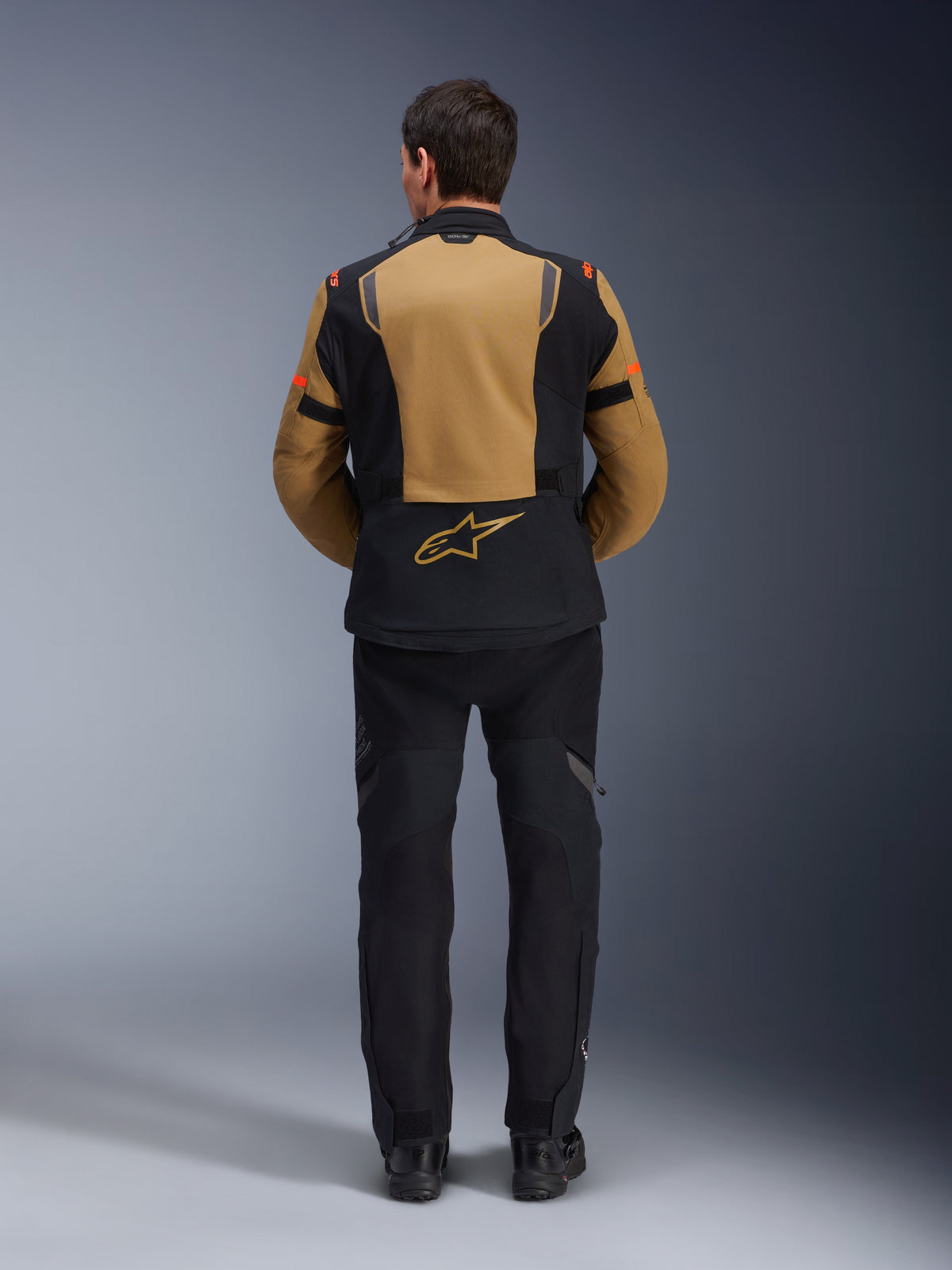 Kurtka Alpinestars ST-7 2L GORE-TEX, przygodowa kurtka motocyklowa, brązowa Gobi i czarna, widok z tyłu prezentujący ergonomiczne panele i logo Alpinestars, zestawiona z czarnymi spodniami i butami motocyklowymi, wodoodporna odzież turystyczna