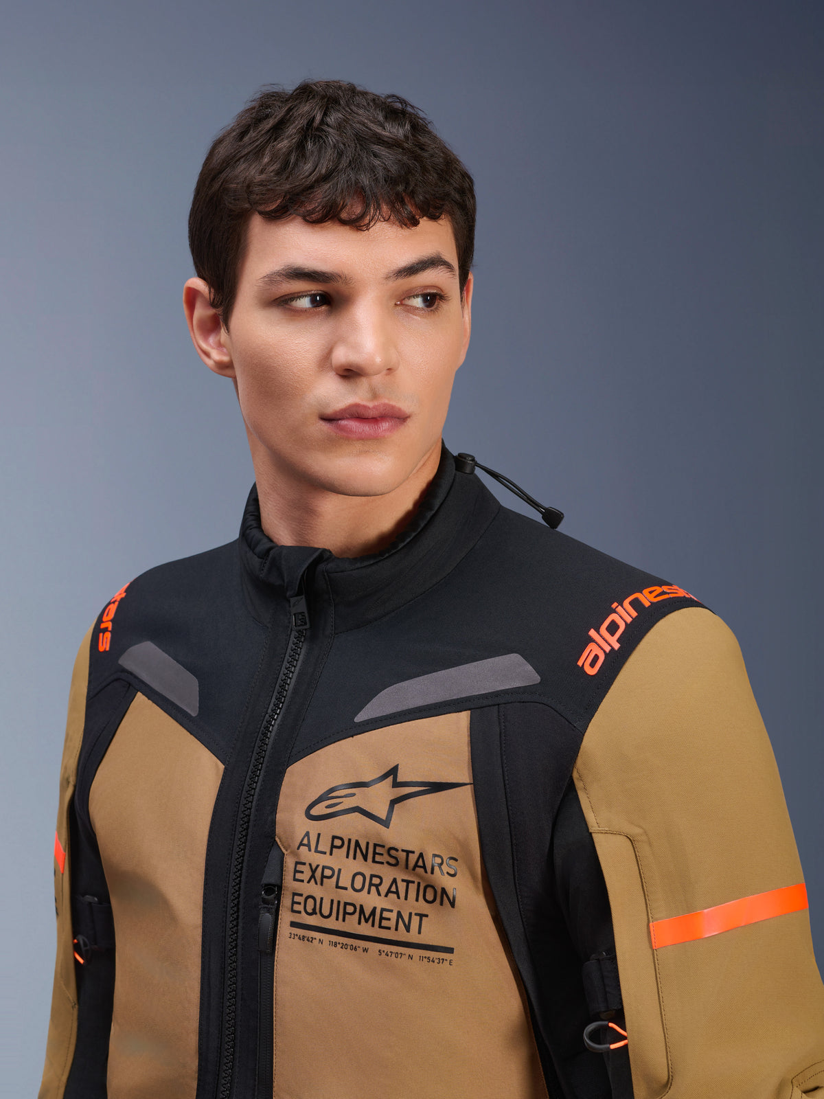 Kurtka Alpinestars ST-7 2L GORE-TEX, kolor brązowy (Gobi Brown) i czarny, prezentowana przez modela w studiu, logo „ALPINESTARS EXPLORATION EQUIPMENT” na klatce piersiowej, pomarańczowe logotypy na ramionach, techniczna wodoodporna konstrukcja tekstylna do jazdy adventure