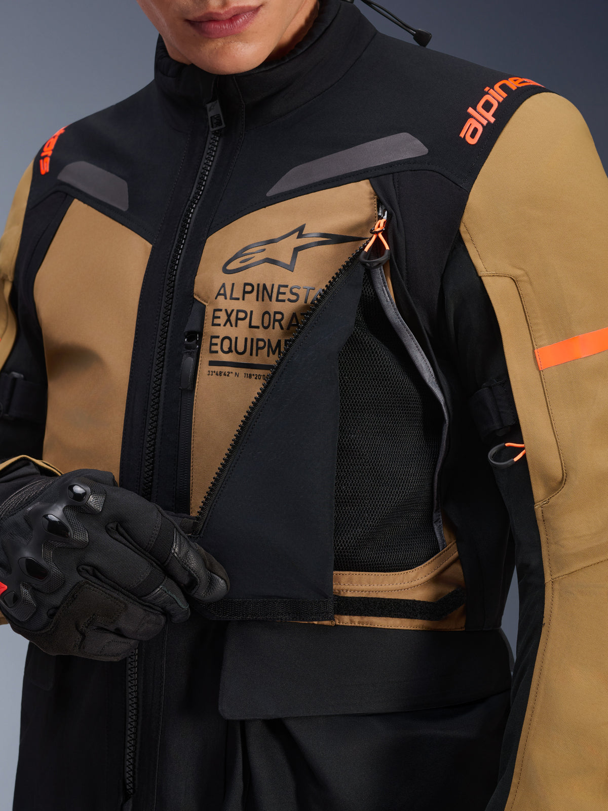 Kurtka Alpinestars ST-7 2L GORE-TEX, motocyklowa kurtka turystyczna adventure, kolor brązowy Gobi Brown i czarny, zbliżenie na rozpięty panel wentylacyjny na klatce piersiowej odsłaniający oddychającą siateczkę, z brandingiem Alpinestars Exploration Equipment i pomarańczowymi akcentami.