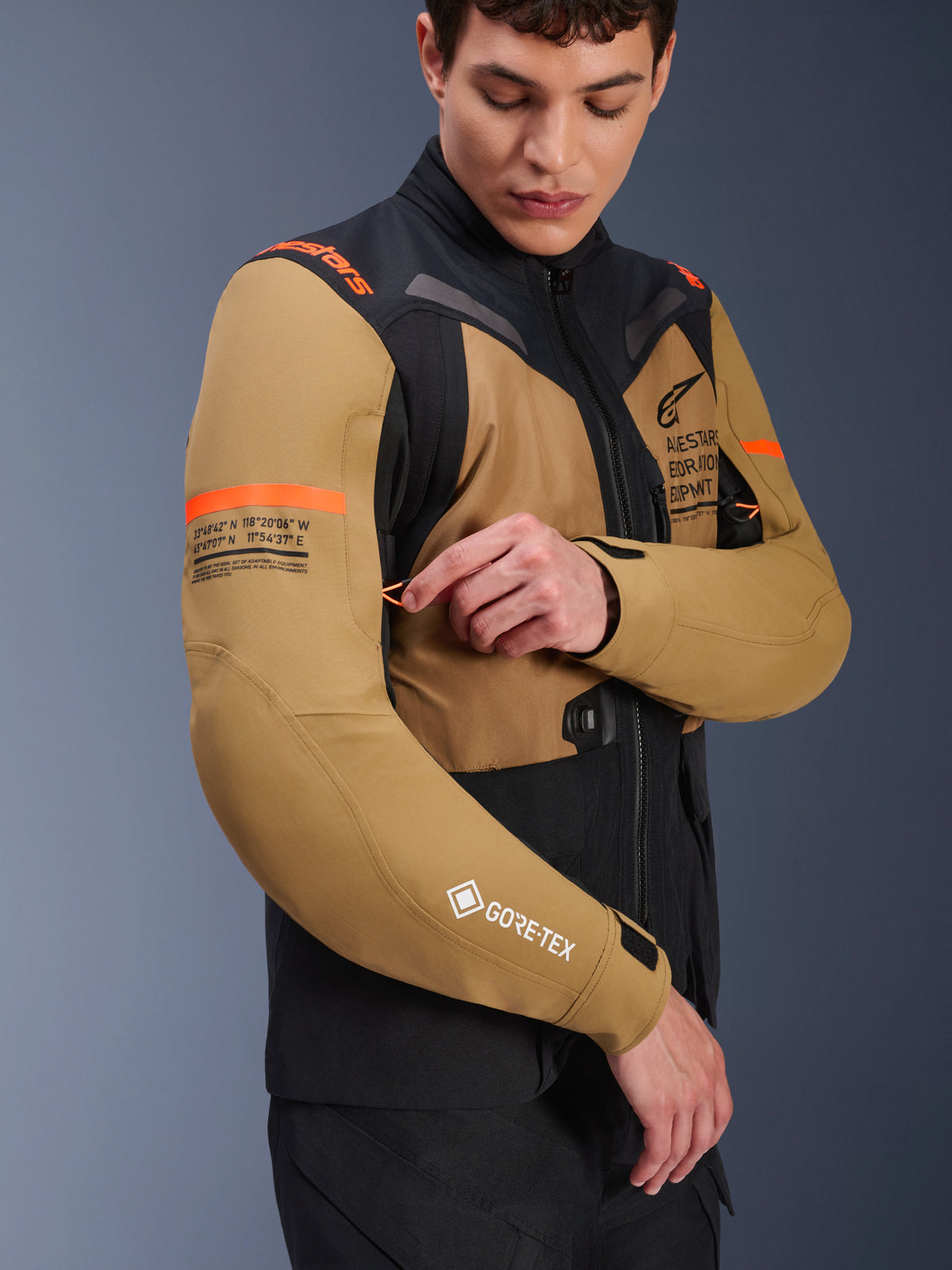 Kurtka Alpinestars ST-7 2L GORE-TEX w kolorze Gobi Brown i czarnym, motocyklowa kurtka adventure-touring noszona przez modela regulującego otwór wentylacyjny na rękawie, pomarańczowe akcenty, grafiki ze współrzędnymi i logo GORE-TEX
