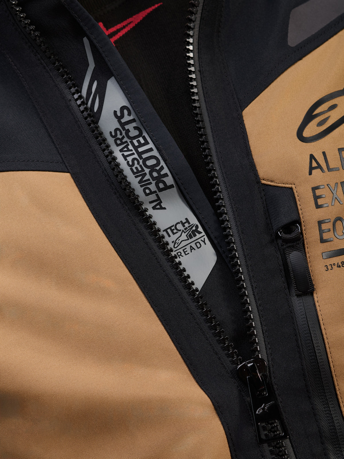 Szczegółowe zbliżenie kurtki Alpinestars ST-7 2L GORE-TEX w kolorze Gobi Brown i czarnym, z brandingiem „ALPINESTARS PROTECTS” i „TECH-AIR READY” wewnątrz głównego zamka, prezentujące wytrzymałą tekstylną konstrukcję oraz logo na klatce piersiowej.
