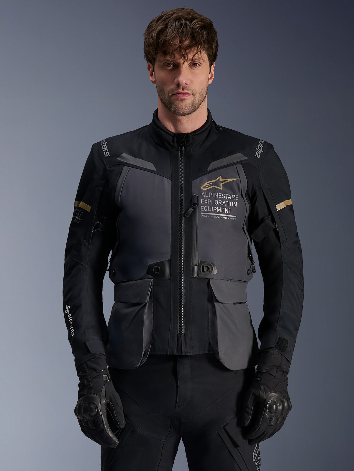 Alpinestars ST-7 2L GORE-TEX Jacket, męska kurtka motocyklowa adventure touring, ciemnoszara i czarna z akcentami khaki, z logo „Alpinestars Exploration Equipment”, wodoodpornymi kieszeniami cargo i wysokim kołnierzem, prezentowana przez modela w czarnych rękawicach