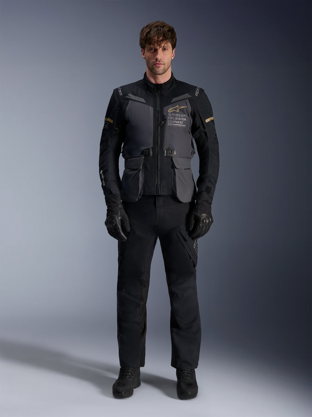Męski model ubrany w kurtkę Alpinestars ST-7 2L GORE-TEX w kolorze Deep Gray Black Dark Khaki, z brandingiem „ALPINESTARS EXPLORATION EQUIPMENT” na klatce piersiowej, w połączeniu z pasującymi czarnymi spodniami motocyklowymi, rękawicami i butami, ujęcie studyjne od przodu.