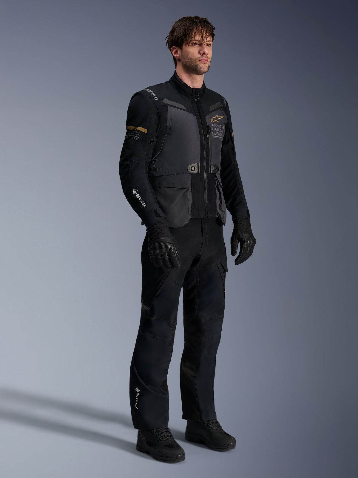 Męski model w kurtce Alpinestars ST-7 2L GORE-TEX w kolorach Deep Gray Black i Dark Khaki, zestaw z czarnymi spodniami i rękawicami adventure, wodoodporna laminowana powłoka z kieszeniami cargo i technicznym brandingiem, idealna do turystyki przygodowej