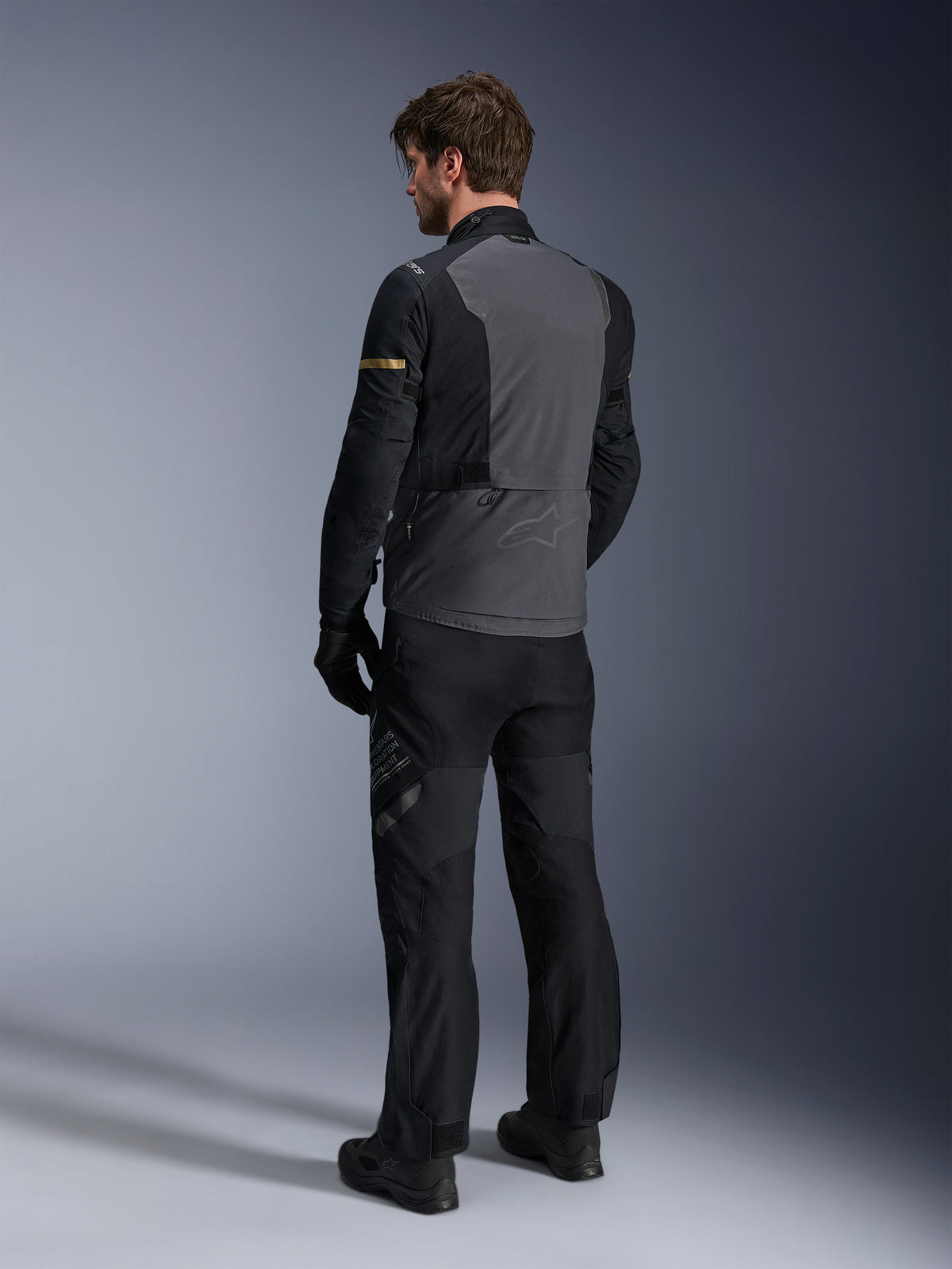 Kurtka Alpinestars ST-7 2L GORE-TEX, ciemnoszara i czarna z akcentami dark khaki, widok od tyłu motocyklisty w kurtce i pasujących spodniach motocyklowych, odzież adventure touring, wodoodporna laminowana konstrukcja