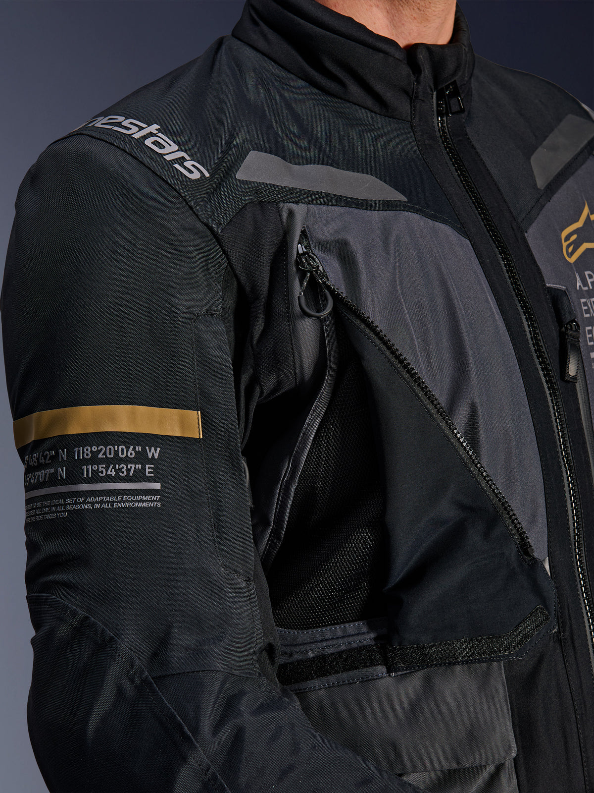 Kurtka Alpinestars ST-7 2L GORE-TEX, ciemnoszara i czarna, zbliżenie na otwarty panel wentylacyjny z siateczką na klatce piersiowej, logo Alpinestars na ramieniu, pasek w kolorze khaki na rękawie z nadrukowanymi współrzędnymi geograficznymi, techniczna odzież motocyklowa adventure-touring