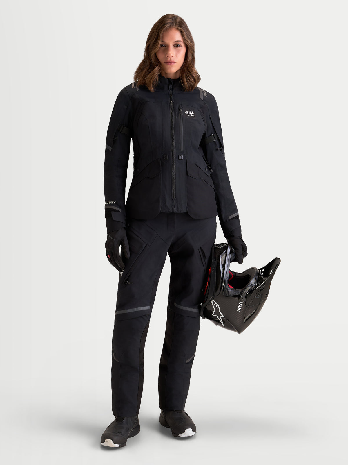 Alpinestars Stella ST-7 V2 2L GORE-TEX damska kurtka motocyklowa adventure w kolorze czarnym, widok na modelce ubranej w dopasowany komplet i trzymającej kask, wodoodporna konstrukcja tekstylna z certyfikowaną ochroną CE