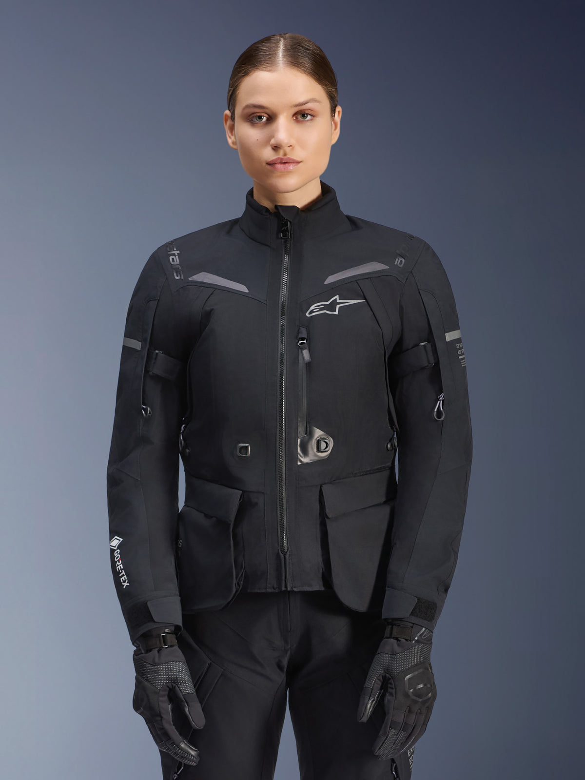 Kobieta ubrana w kurtkę Alpinestars Stella ST-7 2L GORE-TEX, czarna z ciemnoszarymi akcentami, motocyklowa kurtka turystyczna z dużymi kieszeniami cargo i wysokim kołnierzem, zestawiona z czarnymi rękawicami i spodniami, wodoodporna odzież do jazdy adventure