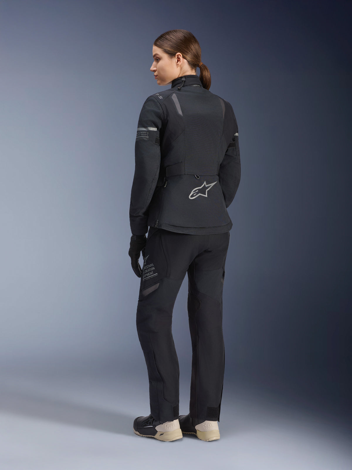 Damska kurtka Stella ST-7 2L GORE-TEX, czarno-ciemnoszara, turystyczna kurtka adventure pokazana z tyłu, noszona z dopasowanymi czarnymi spodniami i kremowymi butami, z logo Alpinestars w dolnej części pleców, zaprojektowana do całorocznej turystyki szosowej.