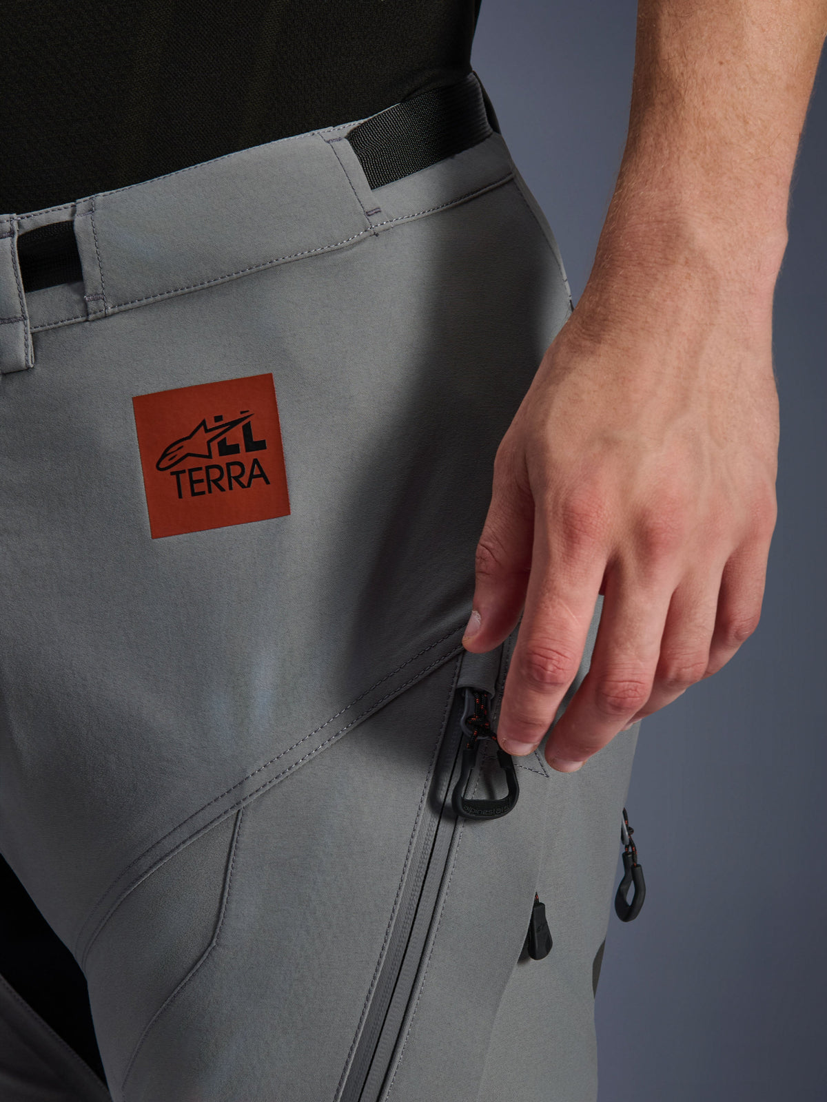 Alpinestars Nazca 3L GORE-TEX PRO Pants, motocyklowe spodnie adventure, ciemnoszare, zbliżenie na górną część uda z rdzawobrązową naszywką z logo All Terra, kieszeń cargo na zamek z czarnymi uchwytami, wytrzymała tekstylna konstrukcja do jazdy w każdym terenie