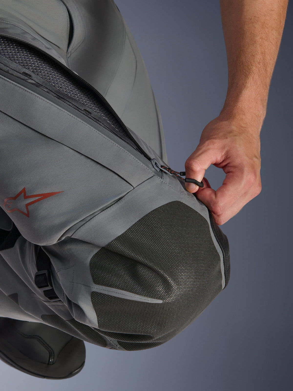 Spodnie Alpinestars Nazca 3L GORE-TEX PRO, motocyklowe spodnie adventure, ciemnoszare i czarne, zbliżenie na dłoń otwierającą zapinany na zamek wlot wentylacyjny na udzie, wzmocnienia kolan z teksturowanego materiału Superfabric, czerwone logo gwiazdy Alpinestars, techniczna konstrukcja tekstylna zapewniająca ochronę w każdych warunkach pogodowych