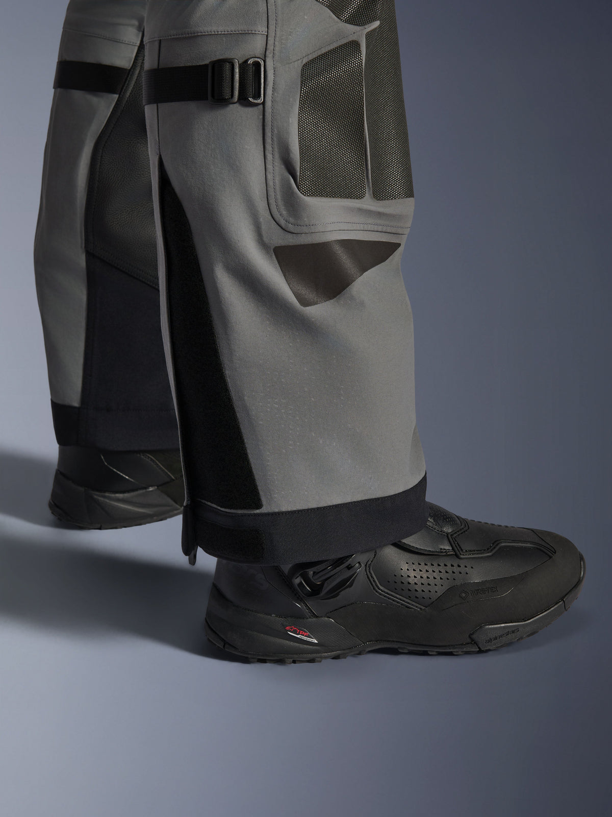 Alpinestars Nazca 3L GORE-TEX PRO Pants, motocyklowe spodnie adventure, ciemnoszare i czarne, zbliżenie na dolną część nogawek z regulatorami objętości łydek i wzmocnieniami Superfabric, noszone z czarnymi butami Alpinestars, wytrzymały materiał tekstylny do ekstremalnej jazdy adventure