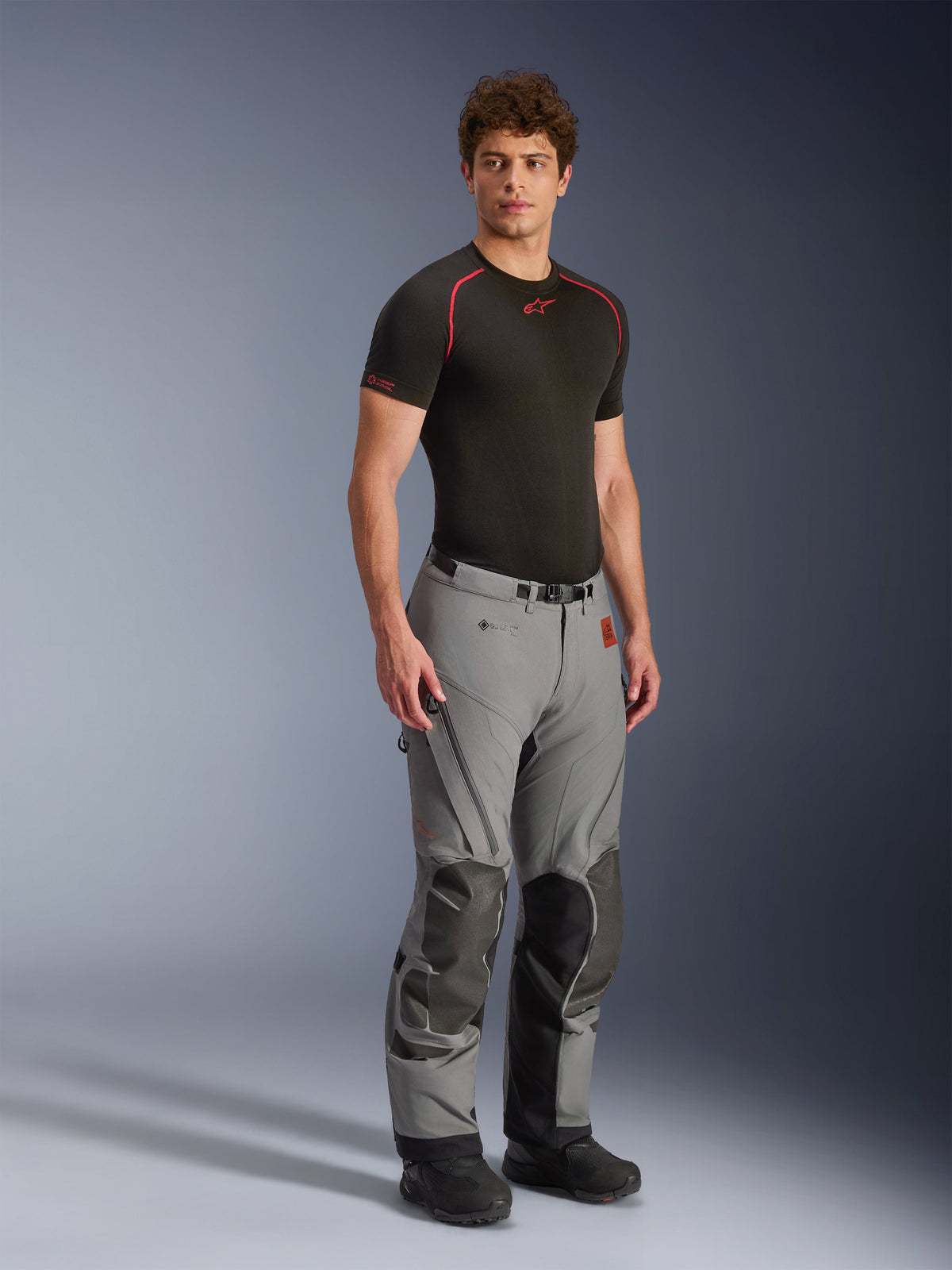 Męski model w spodniach Alpinestars Nazca 3L GORE-TEX PRO, ciemnoszare i czarne spodnie motocyklowe adventure, wzmocnione panele na kolanach i wentylacja na zamek, czarna koszulka termoaktywna, techniczna odzież zaprojektowana do ekstremalnej jazdy w terenie