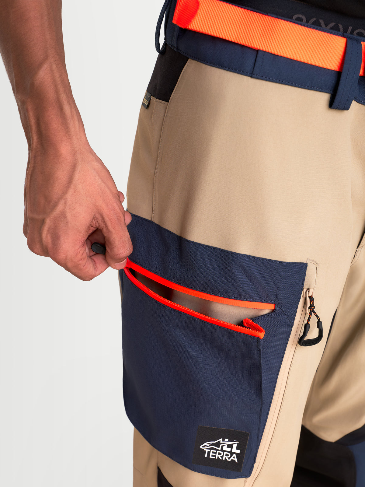 Alpinestars Arenis Gore-Tex spodnie motocyklowe adventure, widok z bliska na rękę otwierającą zintegrowaną kieszeń cargo z jaskrawo pomarańczowymi detalami zamka błyskawicznego, beżowo-granatowa wodoodporna konstrukcja tekstylna