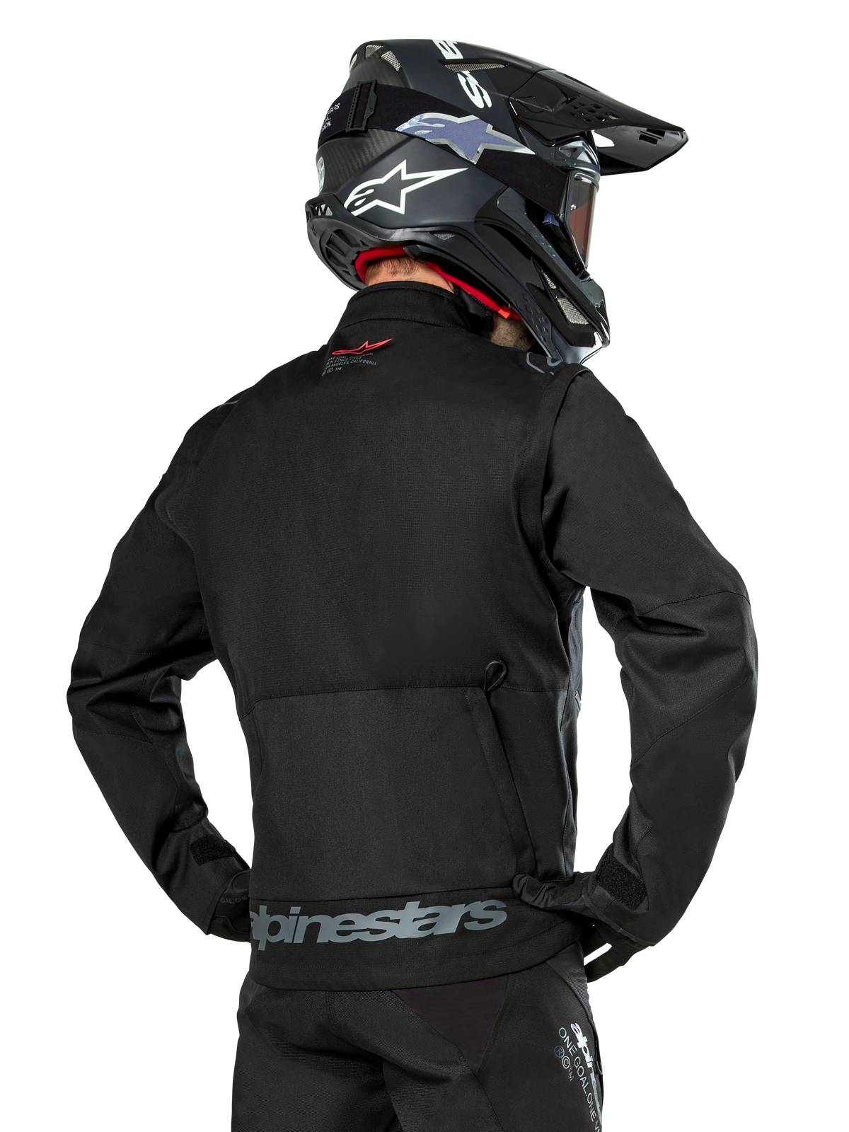 Widok od tyłu motocyklisty ubranego w czarną kurtkę Alpinestars Pro-Dura, wytrzymała tekstylna kurtka enduro z ripstopu, z dużą tylną kieszenią cargo i szarym napisem Alpinestars w dolnej części pleców, w zestawie z czarnym kaskiem motocrossowym.