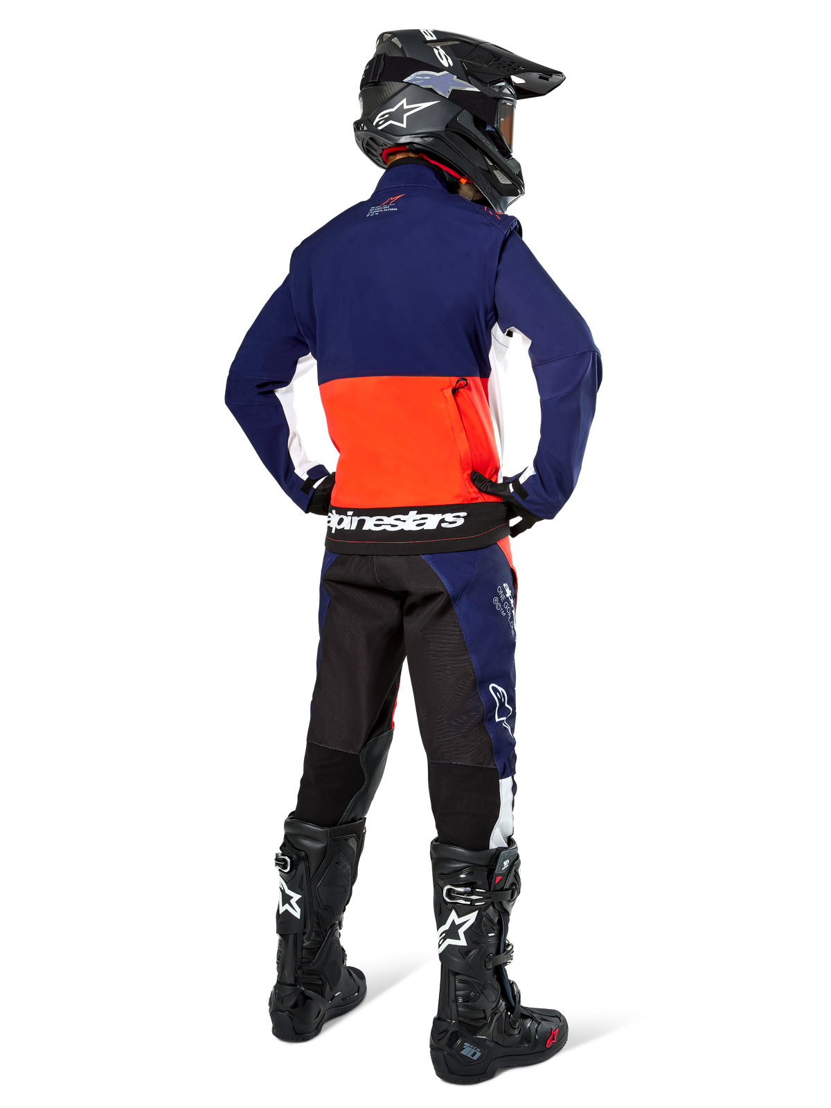 Motocyklista motocrossowy widoczny od tyłu w kurtce Alpinestars Lite-Dura Softshell w kolorach granatowym, intensywnym pomarańczowym i białym, prezentujący dużą tylną kieszeń. Stylizację dopełniają dopasowane spodnie enduro, czarny kask motocrossowy oraz czarne buty off-road.