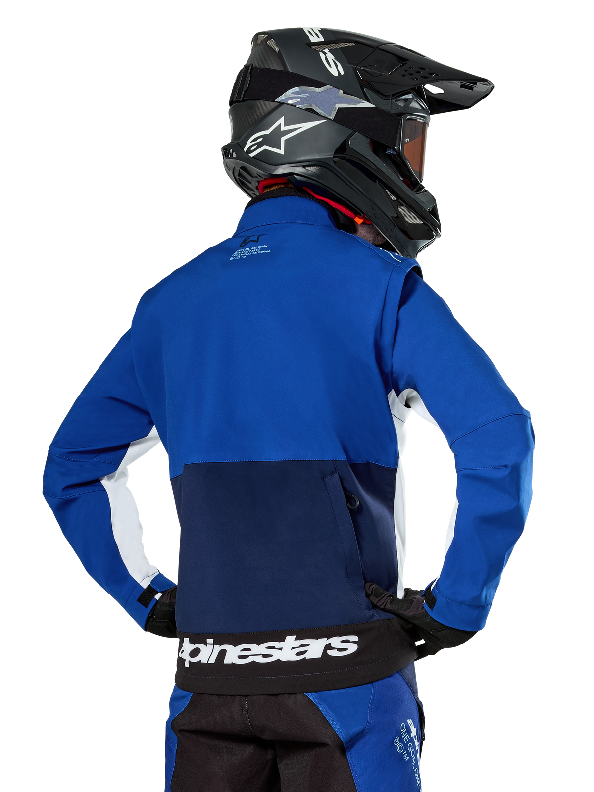Alpinestars Lite-Dura Softshell Jacket in Navy, Bright Blue, and White, rear view showing the large zippered back pocket for sleeve storage, white side accents, and Alpinestars branding on the hem, worn with a black motocross helmet and matching gear.