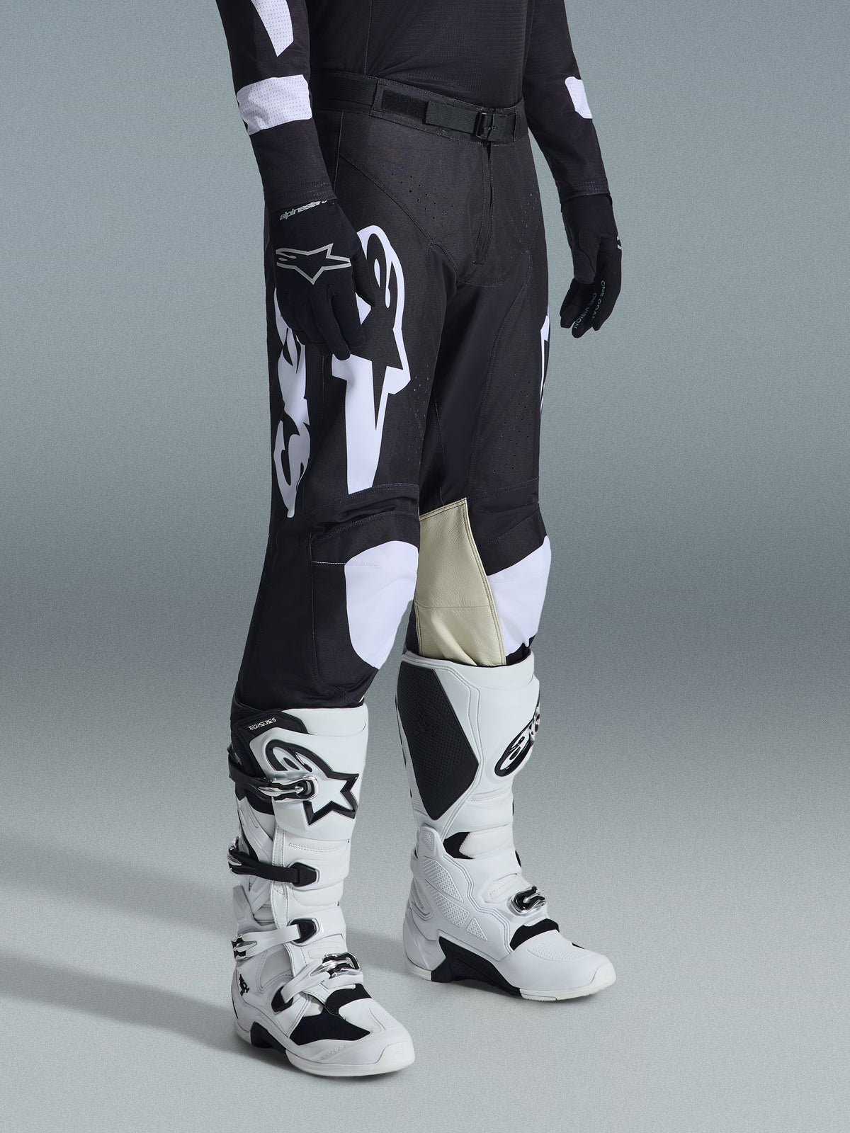 Alpinestars Racer Air Riway Pants 2026, spodnie motocrossowe, czarno-białe, zestawione z biało-czarnymi butami Tech Series i czarną bluzą z długim rękawem, białe logo Alpinestars, laserowo wycinane perforacje oraz jasnobrązowe panele z koziej skóry po wewnętrznej stronie kolan.