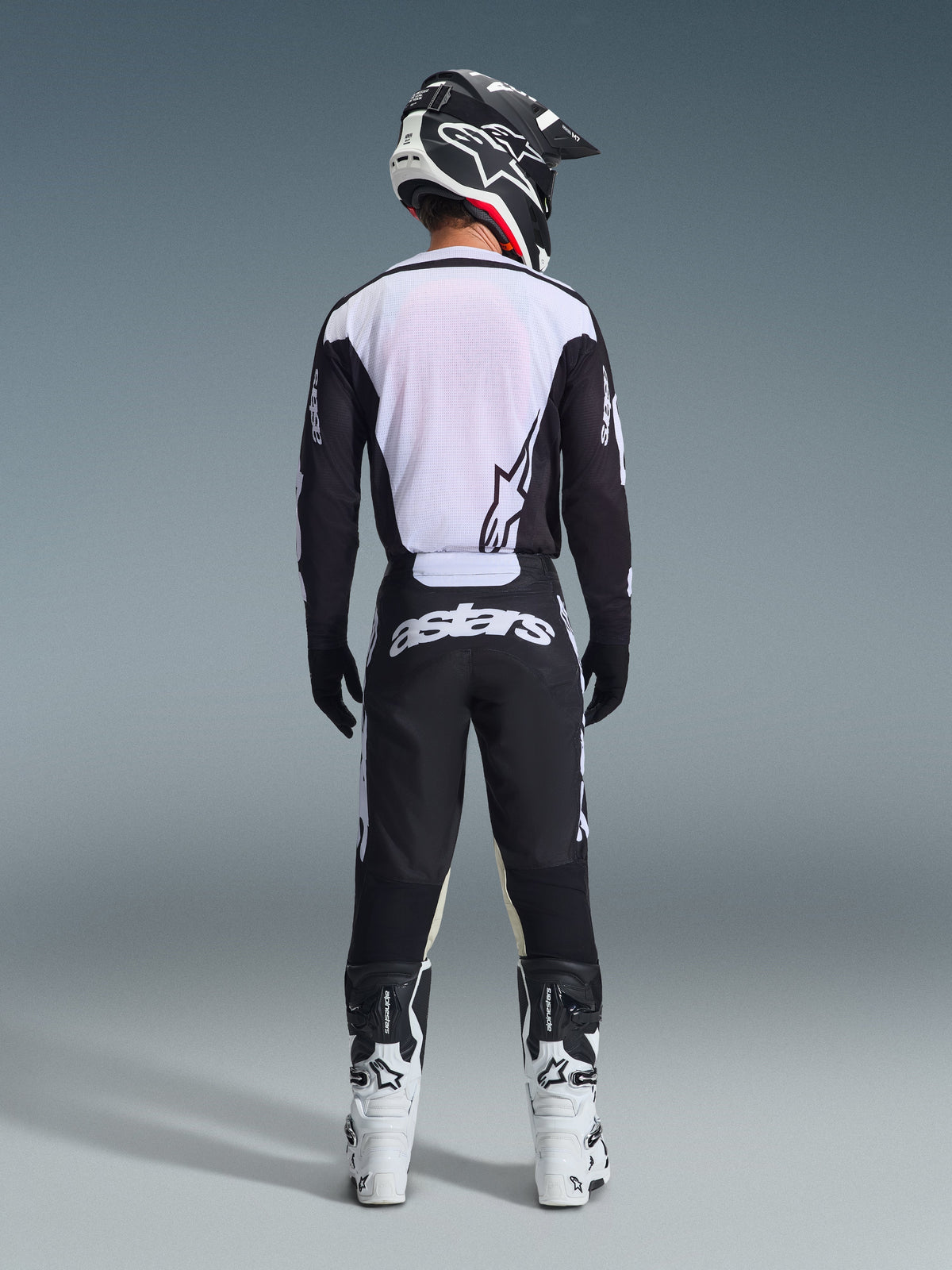 Motocyklista motocrossowy widziany od tyłu w spodniach Alpinestars 2026 Racer Air Riway w kolorze czarno-białym, dopasowanej czarno-białej bluzie z długim rękawem, biało-czarnych butach wyścigowych i czarnym kasku z logo Alpinestars
