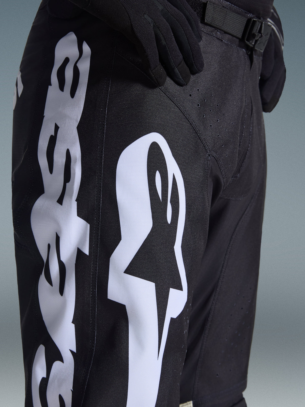 Alpinestars 2026 Racer Air Riway Pants, spodnie motocrossowe, czarno-białe, zbliżenie pokazujące duże białe logo Alpinestars, laserowo wycinane perforacje zapewniające przepływ powietrza oraz regulowaną klamrę w pasie, wytrzymały materiał do jazdy w terenie.