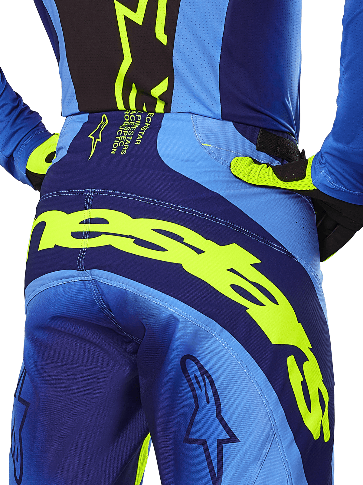 Alpinestars Techstar Melt Pants 2025, spodnie motocrossowe, żółte neonowe i niebieskie, zbliżenie na tył z wyraźnym żółtym napisem Alpinestars na siedzeniu, widoczna niebieska koszulka i żółte neonowe rękawice, profesjonalna odzież do wyścigów terenowych