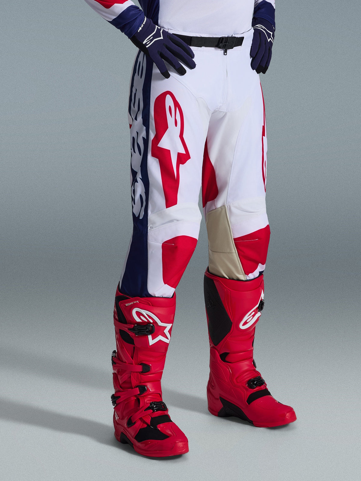Alpinestars Racer Riway Pants 2026, spodnie motocrossowe, biało-niebiesko-czerwone, z wyraźnymi czerwonymi logo na udach i niebieskimi panelami bocznymi z białym brandingiem, noszone z czerwonymi butami i ciemnoniebieskimi rękawicami, ergonomiczna konstrukcja do wyścigów off-road