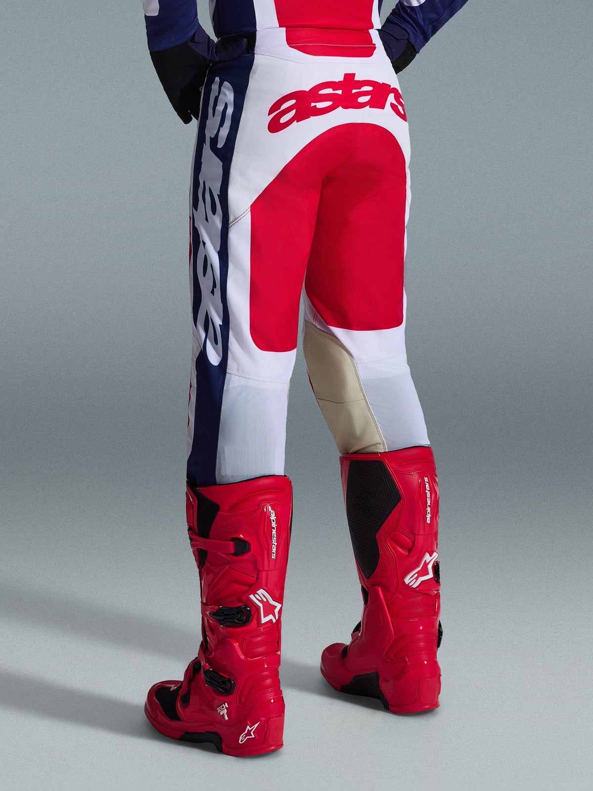 Alpinestars 2026 Racer Riway Pants, spodnie motocrossowe, biało-niebiesko-czerwone, widok z tyłu przedstawiający duże czerwone logo Astars na siedzeniu i beżowe skórzane panele na wewnętrznej stronie kolan, noszone z czerwonymi butami motocrossowymi Alpinestars, profesjonalna odzież do jazdy terenowej