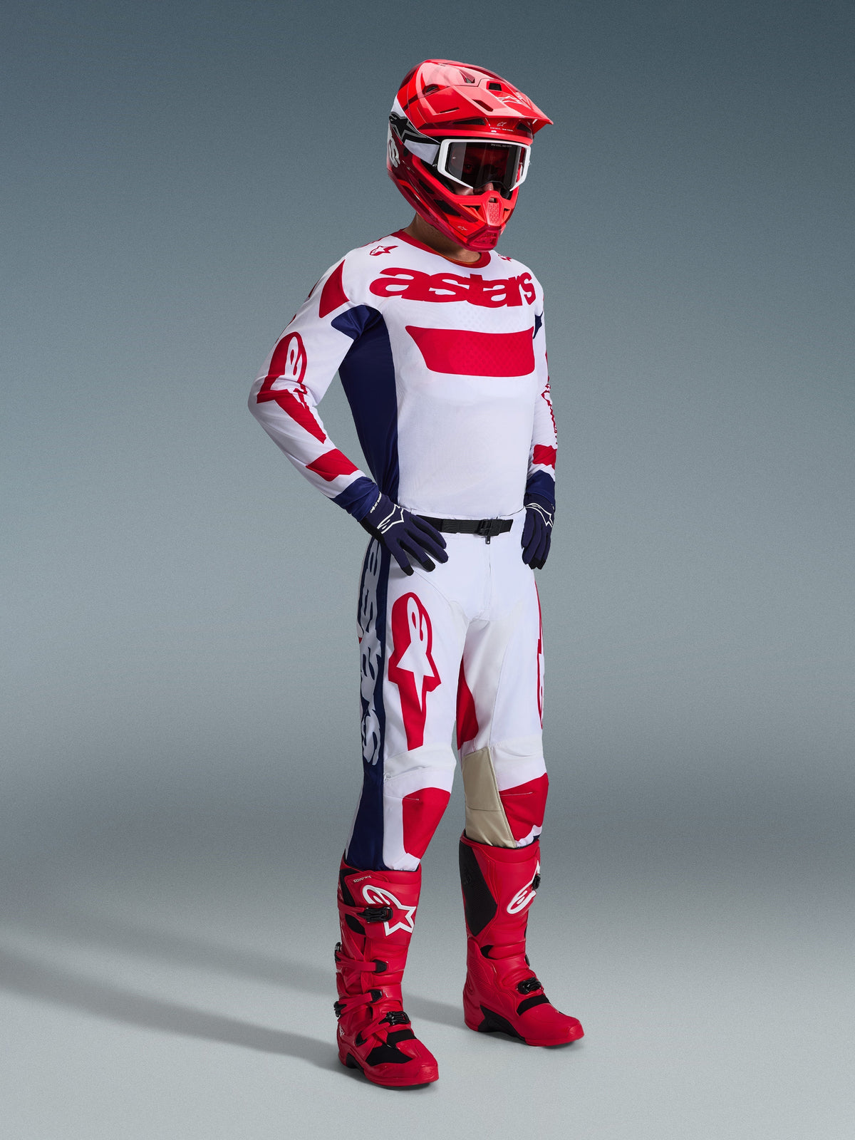 Zawodnik motocrossowy w pełnym stroju wyścigowym, spodnie Alpinestars 2026 Racer Riway w kolorystyce białej, niebieskiej i czerwonej, charakteryzujące się białą bazą z wyrazistymi czerwonymi logo i niebieskimi panelami bocznymi, w zestawie z pasującą biało-czerwoną koszulką, czerwonym kaskiem i czerwonymi butami motocrossowymi.