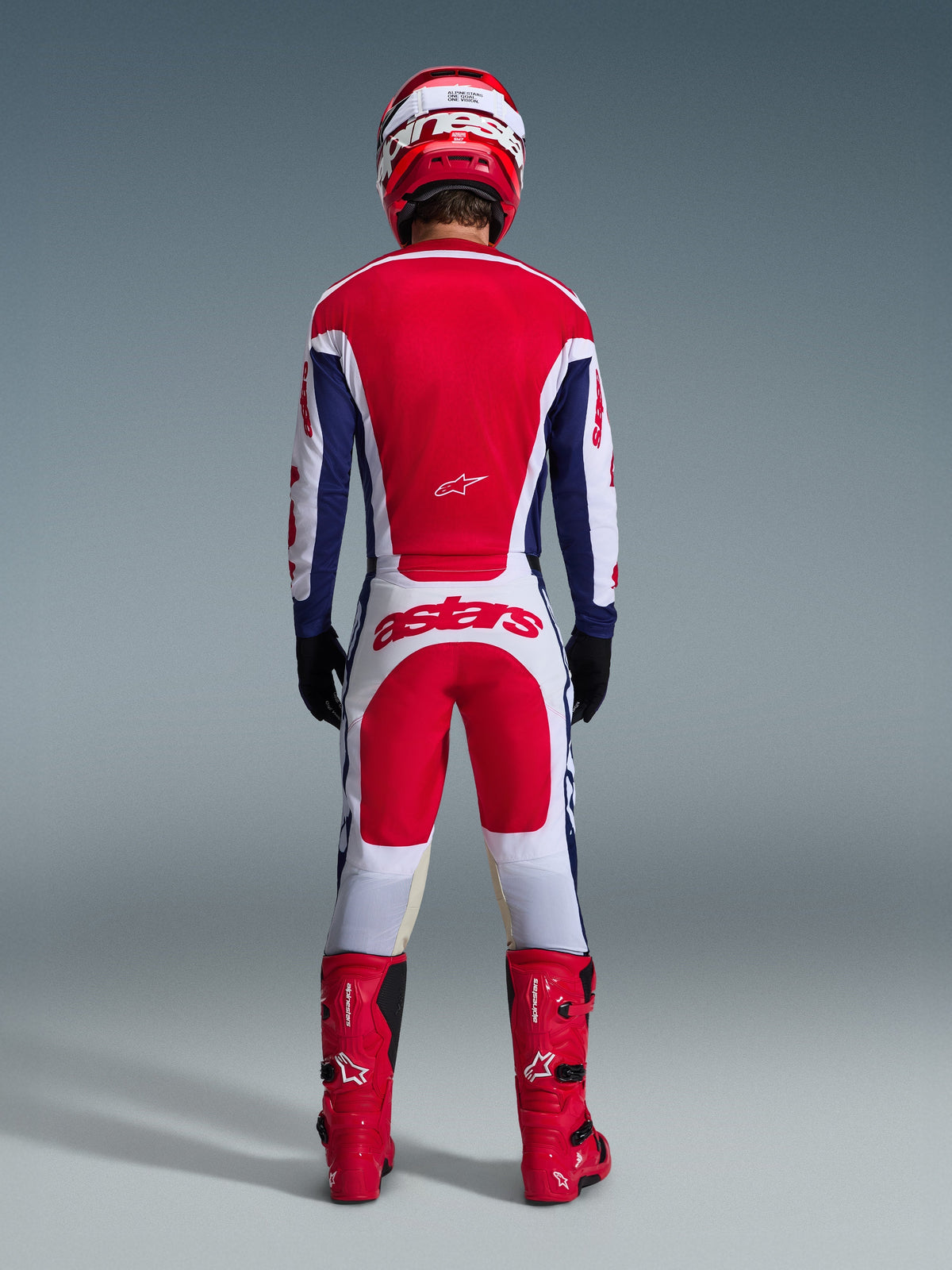 Motocyklista motocrossowy od tyłu w pełnym stroju wyścigowym, spodnie Alpinestars 2026 Racer Riway w kolorze biało-niebiesko-czerwonym z pasującą bluzą, czerwone buty i kask, duże logo „astars” na siedzeniu, spójny strój motocrossowy.