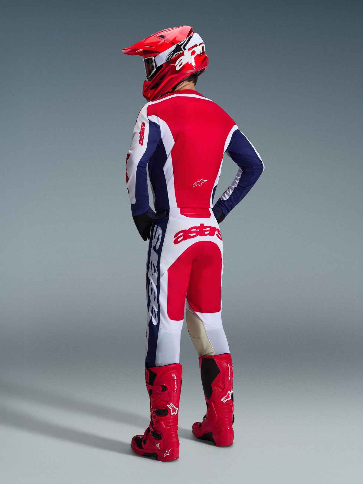 Zawodnik motocrossowy widziany od tyłu, ubrany w spodnie Alpinestars 2026 Racer Riway w kolorach białym, niebieskim i czerwonym, w zestawie z pasującą koszulką, czerwonym kaskiem i jaskrawoczerwonymi butami, prezentujący wyraźne logo na siedzeniu i sportowy krój off-road.