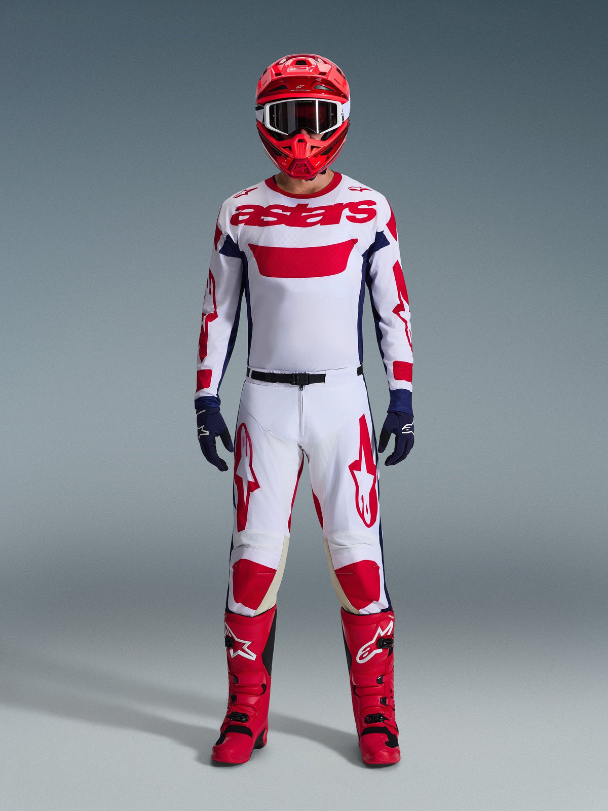 Motocyklista motocrossowy w kompletnym stroju wyścigowym, biała koszulka z czerwonym brandingiem Alpinestars i niebieskimi akcentami, spodnie Alpinestars 2026 Racer Riway w kolorystyce White Blue Red, czerwony kask z dopasowanymi goglami, czerwono-czarne buty, granatowe rękawice, skoordynowany zestaw do wyścigów terenowych